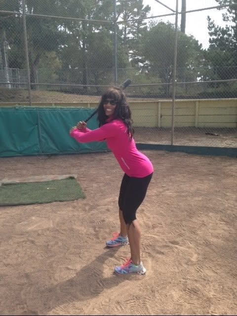 Chilone in a magenta top holding a baseball bat at a batting cage, looking back and smiling